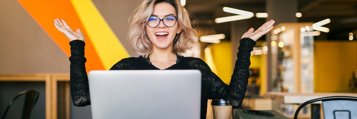 funny happy excited young pretty woman sitting at table in black shirt working on laptop in co-working office, wearing glasses